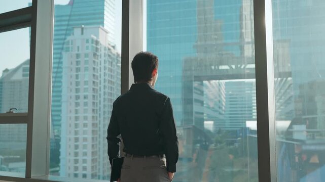 Young Asian businessman looking out the window at a modern city skyline from a high-rise office, symbolizing confidence, ambition, leadership, and future business vision