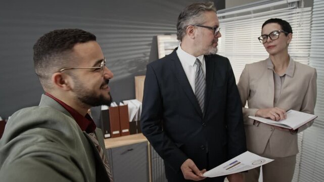 POV of carefree zoomer smiling at camera recording social media video with two elegantly dressed mature coworkers looking at him disapprovingly in office