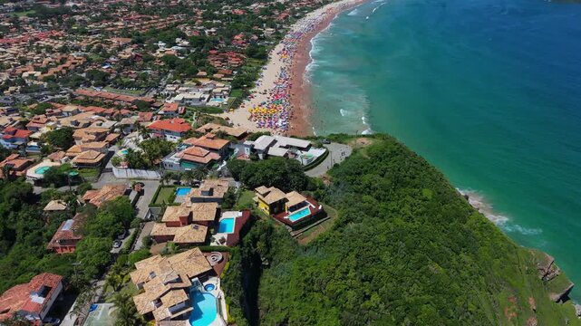 Sweeping aerial drone footage revealing the massive, crowded white sands of Praia de Geriba in Buzios. Colorful umbrellas dot the beach next to the vibrant blue ocean.