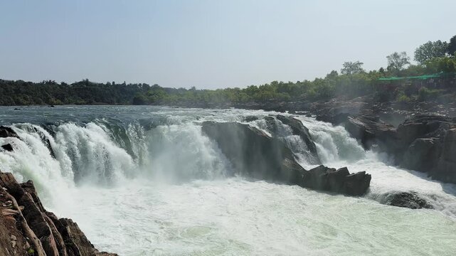 Wide shot of a powerful waterfall cascading over rocky cliffs with foaming water and mist rising above the river, surrounded by green forest landscape.