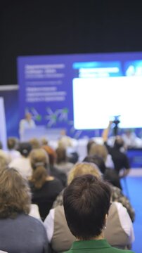 Large group of people are sitting in a room and watching a presentation. There are several people in the front row and many more in the back