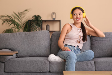 Obraz premium Beautiful young happy Asian woman in headphones listening music on sofa at home