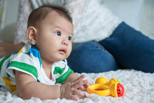 Cute Asian baby boy in striped shirt lying on tummy on white blanket looking away. Little infant playing with yellow toy rattle on bed with mother in blurred background. Parenting and care concept.
