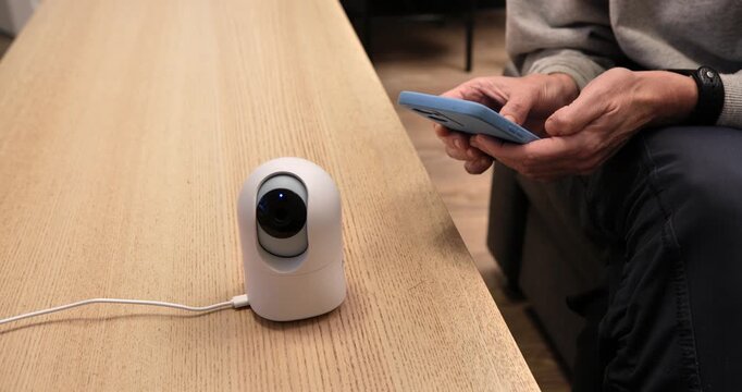 Smart Home Security: Close-up of a compact, white smart security camera on a desk, connected to a wire, with a man using a phone to remotely manage his home's safety.