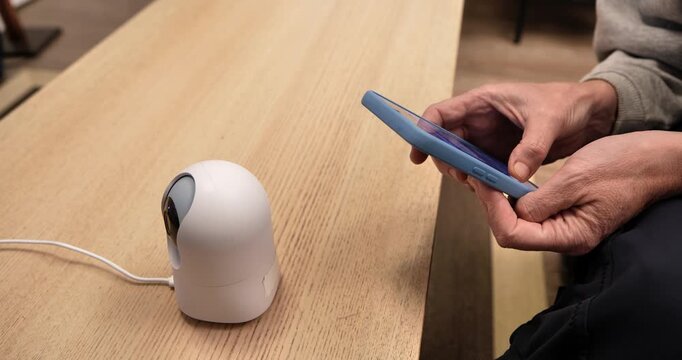  Smart Home Innovation: An individual interacts with a smartphone alongside a sleek, modern security camera, symbolizing the fusion of technology and home security. 