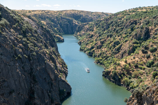 Vista panor&acirc;mica do canh&atilde;o do Rio Douro em Miranda do Douro, com encostas rochosas e escarpadas cobertas de vegeta&ccedil;&atilde;o  nas margens, onde um barco tur&iacute;stico navega tranquilamente sob um c&eacute;u claro