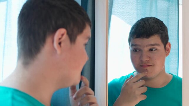 A teenager applies acne cream to problem skin on his face while looking in a mirror at home.