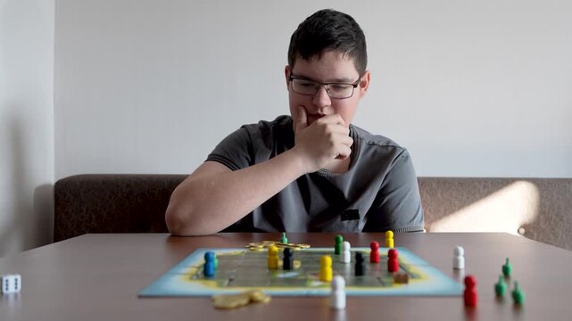 Dice and game pieces sit on a table while a teenager in the background plays a board game, moving pieces across the board.