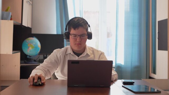 A teenager in glasses sits at a desk in a room during the day, thoughtfully gazing at the screen. a gamer plays on a computer