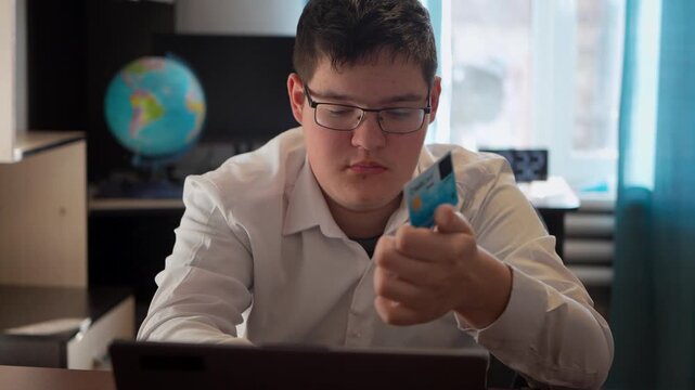 A teenage boy holds a bank card in his hand while sitting at a laptop. the guy enters the card details on the website.