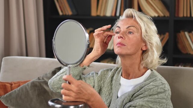 Love yourself. Beautiful old mature woman enjoying mirror reflection. Senior adult older lady looking at mirror confident happy on sofa at home. Woman doing daily morning beauty routine. Self love