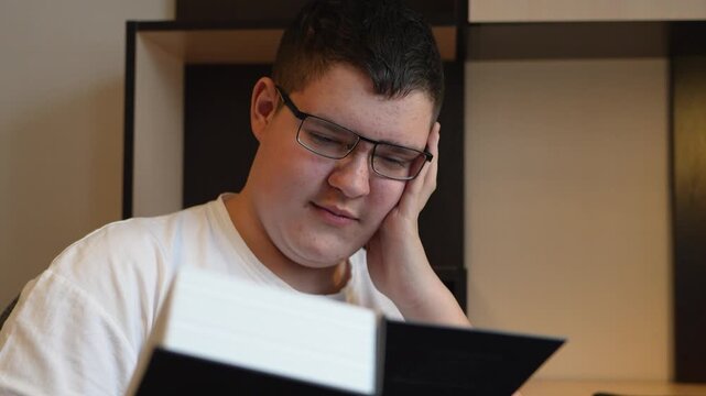 Reading book and doing homework. A teenage boy with glasses reads a book lying on the couch during the day.
