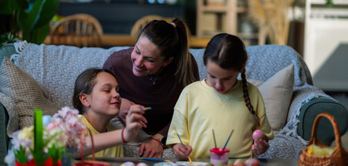 Obraz premium Loving mother watching her two happy daughters painting colorful easter eggs together at home, sharing a special moment during spring holiday preparations with family closeness