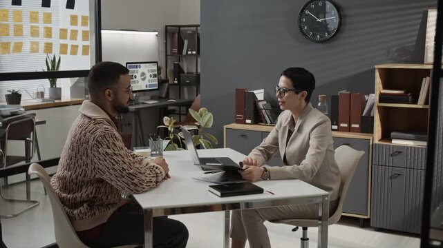 Mature female HR specialist wearing pantsuit telling gen Z male candidate about working conditions while holding job interview at white table in office space