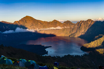 Naklejka premium Colorful tents perched on a high mountain ridge above a vast crater lake at sunrise.