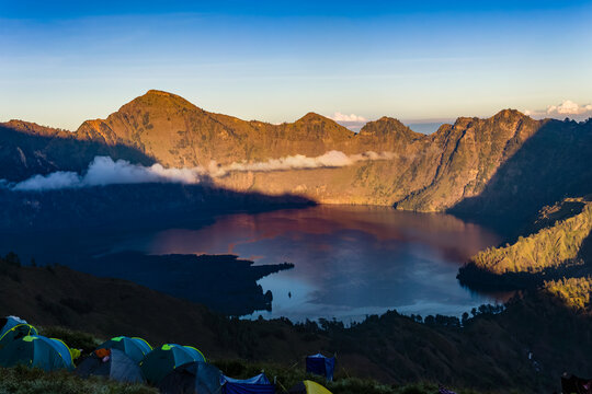 Colorful tents perched on a high mountain ridge above a vast crater lake at sunrise.