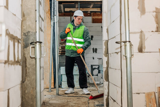 Builder wearing hard hat and vest sweeping construction site indoors