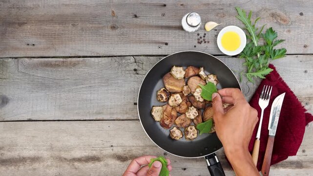 A frying pan with fried potatoes and zucchini sits on an old wooden table. Men's hands are adding spinach leaves to the dish. Parsley, arugula, a clove of garlic, vegetable oil are on the table.