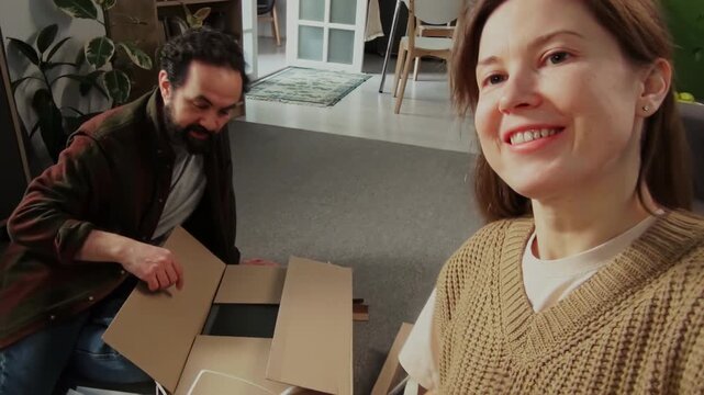 Handheld medium zoom shot of cheerful Caucasian woman filming on digital camcorder herself and husband unpacking parts of DIY assembly cabinet at home, demonstrating pieces, for family memory archive