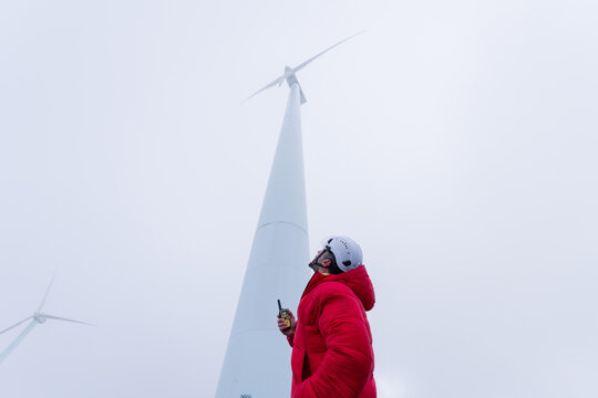 Technician in red jacket inspecting wind turbine outdoors on cloudy day