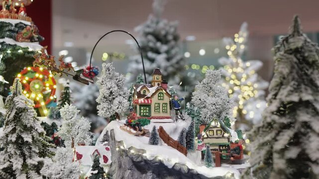 A close-up of an animated miniature Christmas village. Santa leigh and reindeer fly over snowy houses and trees in a magical, illuminated diorama. Festive holiday decoration in motion