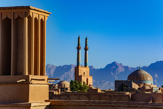 Sunlit Yazd skyline in Iran featuring a tiled mosque dome, twin minarets and a traditional windcatcher (badgir) rising above adobe rooftops with rugged mountain ranges framing the clear blue sky.