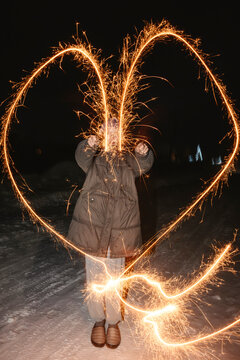 Person in warm clothes drawing heart shape with sparklers at night in winter