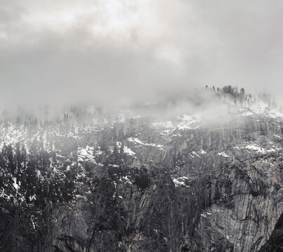View of a rugged mountain face shrouded in ethereal mist and cloud cover, its slopes dotted with trees and snow patches, creating a monochrome landscape, California, United States.