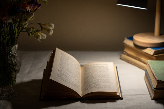 Open book under warm desk lamp light with flowers and stack of books, cozy evening reading still life.