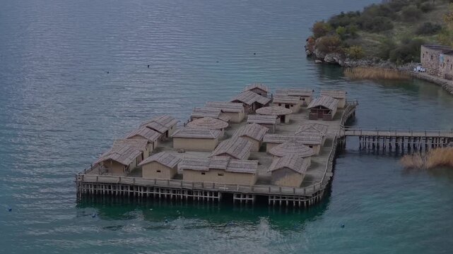 A telephoto drone orbit of the prehistoric Bay of Bones Museum on Lake Ohrid, showing the detailed textures of the wooden houses and thatched roofs against the deep blue water.