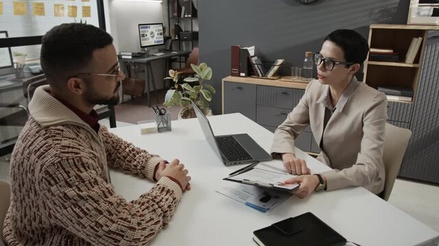 Middle-aged female staff coordinator with short hair commenting on resume of young male applicant who sitting at office table opposite her