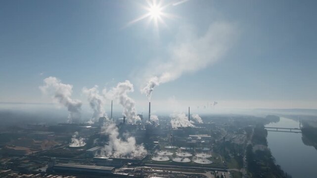 High drone view showcasing multiple chimneys releasing steam over a large factory footprint, with industrial basins and rooftops visible under bright daylight.