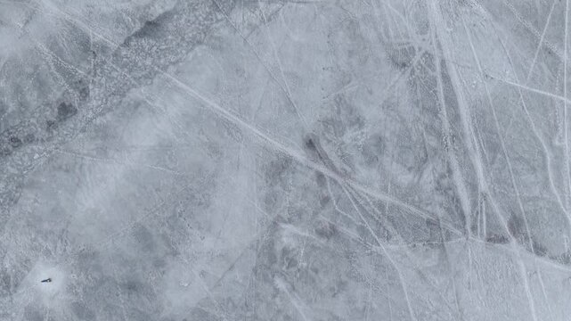 Aerial footage capturing a frozen lake with intricate skating marks from above. The icy texture and patterns create a mesmerizing winter landscape under soft, diffused lighting.