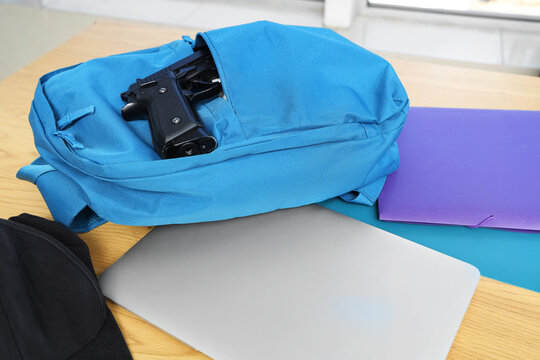 Gun with backpack, laptop and copybooks on desk in classroom, closeup. School shooting concept
