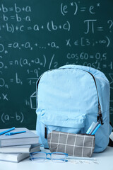Naklejka premium Blue school backpack with eyeglasses and stationery on white table near green chalkboard