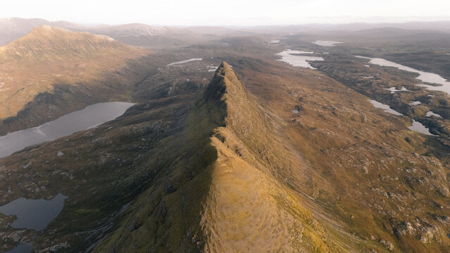 Aerial view of the rugged, sharply defined peak of Suilven Mountain cuts through a vast, undulating landscape speckled with lochs, Assynt, United Kingdom.