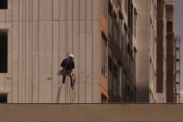 Construction worker wearing safety gear is suspended side of tall building, performing maintenance or cleaning tasks. urban setting and high rise architecture create sense of height and scale © pirom