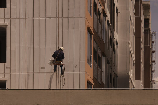 Construction worker wearing safety gear is suspended side of tall building, performing maintenance or cleaning tasks. urban setting and high rise architecture create sense of height and scale