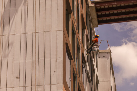 Construction worker wearing safety gear is suspended side of tall building, performing maintenance or cleaning tasks. building features modern design with large windows and concrete panels clear