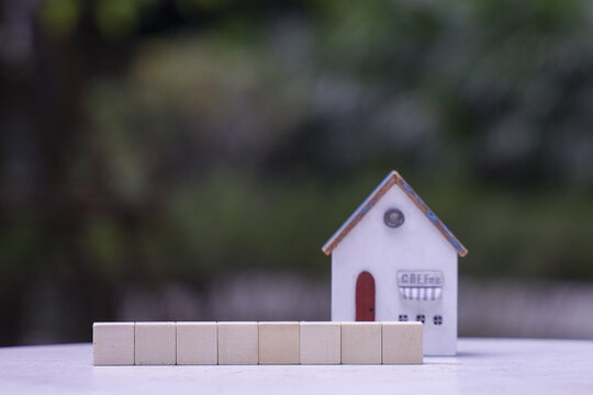 Small model house with red door and sign reading Cafe stands surface, surrounded by blurred green background. In front of house, there are several wooden blocks aligned row, creating minimalist