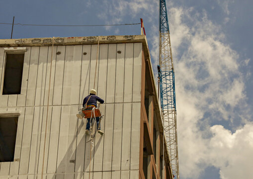 Construction worker wearing safety gear is suspended side of building, performing maintenance or installation work. scene captures height and scale of construction site against partly cloudy sky