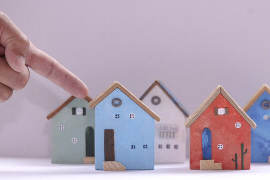 Miniature colorful wooden houses with hand pointing at blue one, creating playful and whimsical scene. houses are arranged in row, each with unique details and colors