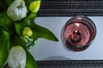 A glass of wine placed on a white countertop, the neck of a wine bottle from which wine is poured...