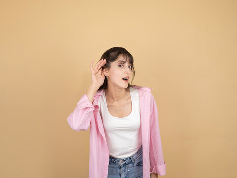 A curious young woman in a pink shirt cups her hand to her ear, leaning forward to listen intently. She stands against a neutral beige background with a surprised and inquisitive facial expression.