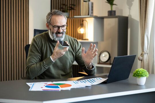 Man using voice assistant on phone while working on laptop with business charts on desk, communicating remotely