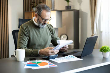 Obraz premium Mature man working in a home office, calculating expenses and reviewing financial statistics and paperwork on his desk