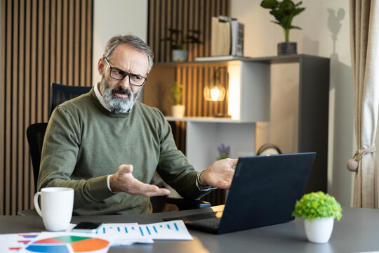 Mature man feeling frustrated and stressed, gesturing confusedly while looking at a laptop computer in a contemporary home office