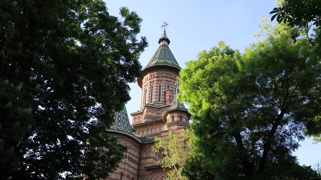 Ornate brick dome of an orthodox cathedral framed by lush green trees, Timisoara, Romania