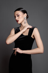 Naklejka premium Beauty fashion woman stands in studio with a black outfit. She poses with her hand near her neck. Her hair is styled back, and she wears large earrings. lighting is soft, focusing on her features