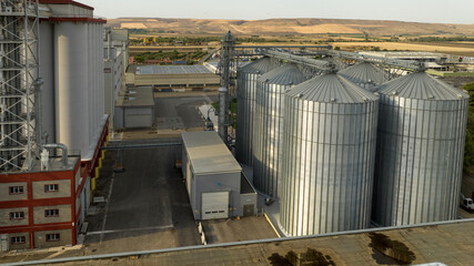 Aerial view of a massive industrial grain facility featuring towering silver metal silos and white storage bins. The complex includes elevator towers and conveyors set against a rural landscape. © Stefano Tammaro